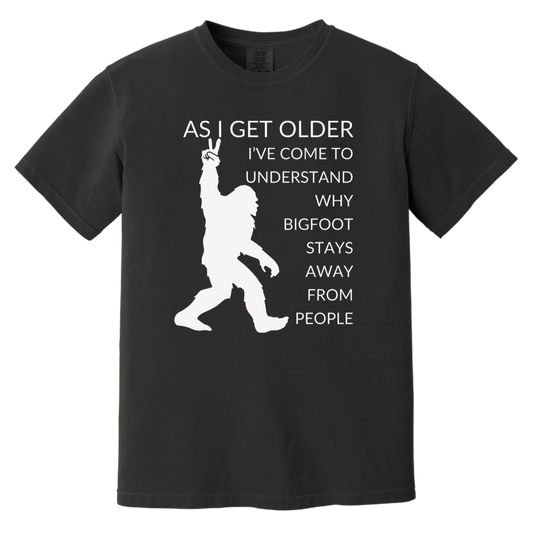 Black Heavyweight Garment-Dyed Cotton T-Shirt featuring the inspiring quote, "As I get older, I've come to understand why Bigfoot stays away from people," uniquely formatted along side a silhouette of Bigfoot. This t-shirt combines comfort and style, making it a perfect addition to your wardrobe.