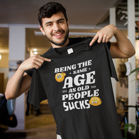 Black heavyweight garment-dyed t-shirt featuring the humorous quote "Being the same age as old people sucks" with colorful emoji graphics.