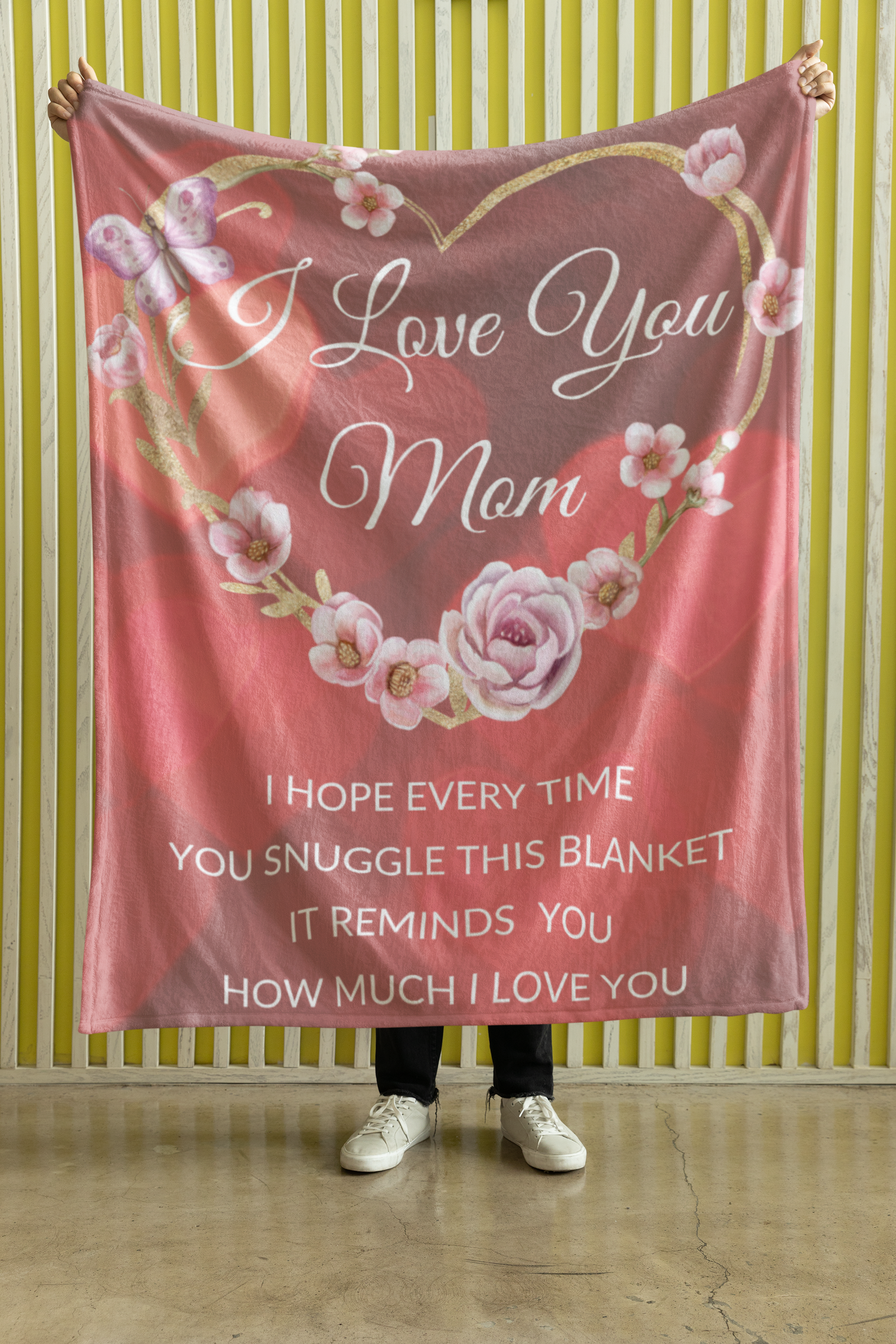 A premium Sherpa blanket with a design of a flower-covered golden heart and the message 'I love you mom'. The blanket offers ultimate comfort with a silky smooth front, fluffy sherpa back, a luxurious thickness of 1 cm. held up by someone to showcase the design